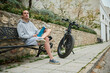 © Taras Grebinets - Young Caucasian man in gray hoodie and sports shorts, sitting on a bench near his rented electric bike, relaxing in the urban environment