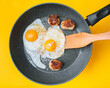 Fried eggs in a frying pan next to three cutlets on a yellow background. Bachelor breakfast, lunch and dinner concept.