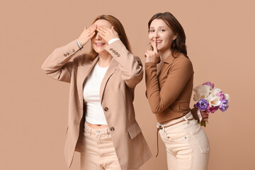 Wall Mural - Beautiful mature woman and her daughter with bouquet of tulips showing silence gesture on brown background. Mother's Day celebration