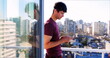 Man in purple shirt standing on balcony using smartphone, cityscape in background