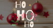 Two red Christmas ornaments hang in front of festive backdrop with HO HO HO text