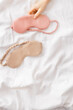 Close up two eye masks for sleep beige and pink color on white bedclothes, woman hand take one, minimal flat lay. Top view sleeping mask for best sleeper, comfort relax. Rest well concept.