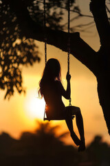 Wall Mural - A silhouette of a girl sitting on a tree swing at sunset creates a backlit image of a young woman swinging in the park.