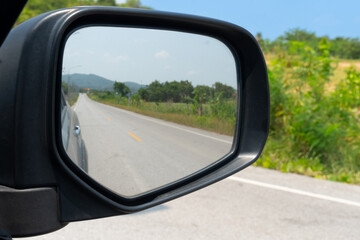 Poster - View of mirrors wing. Rear view of a gray car with asphalt road and green trees and bright sunlight at daytime. Beside road with blurred yellow flower and tree under blue sky.