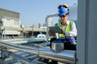 © ultramansk - Young technician with hard hat and safety vest adjusts equipment on a commercial building's rooftop amidst an urban landscape under a clear sky.