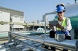 © ultramansk - Young technician with hard hat and safety vest adjusts equipment on a commercial building's rooftop amidst an urban landscape under a clear sky.