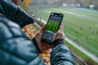 © Vovmar - A man with a mobile phone in his hands watches an online football broadcast at the stadium. Concept of sports applications on mobile devices. Generated by artificial intelligence