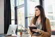 © Bliss - A woman stands before a computer, holding a clipboard, engrossed in her work within a bustling office atmosphere.