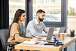 © Bliss - Two professionals sit at a table in an office, engaged in work on their laptops, showcasing a modern corporate culture.