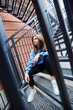 A student girl is sitting in beautiful clothes on an iron staircase. A photo shoot for a girl.