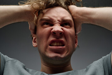 Wall Mural - Low angle closeup studio portrait of young Caucasian man expressing feeling of wrath, gray background