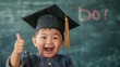 A joyful child wearing a graduation cap gives a thumbs up in front of a chalkboard