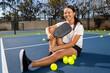 © Mat Hayward - Young female pickleball player sitting outside on the court. She is holding paddle or racquet and surrounded by yellow game balls.