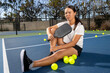 © Mat Hayward - Young female pickleball player sitting outside on the court. She is holding paddle or racquet and surrounded by yellow game balls.