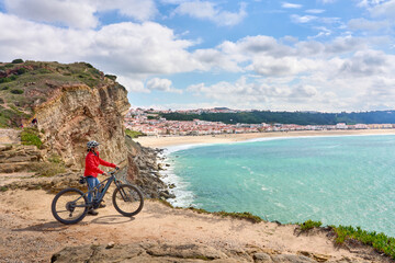 Wall Mural - nice senior woman riding her electric mountain bike on the rocky cliffs above the city of Nazare at the western atlantic coast of Portugal