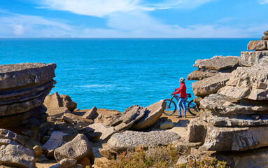 Wall Mural - Brave senior woman riding her electric mountain bike on the rocky cliffs of Peniche at the western atlantic coast of Portugal