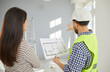 © Studio Romantic - Back view from behind professional architect, engineer or builder in yellow uniform vest and white hard hat holding sheet of paper with house construction blueprint plan and talking to woman homeowner