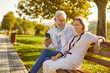 Smiling happy senior active couple man and woman sitting on the bench in a public park using mobile phone together and laughing outdoors. Technology and elderly retired people concept.