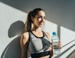 © Monika - Młoda kobieta w sportowym stroju oświetlona promieniami słońca stoi na tle ściany trzymając w ręku butelkę z wodą. Lato, sport, wakacje, zdrowy styl życia
