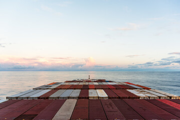 Wall Mural - View on the containers loaded on deck of the large cargo ship. She is sailing through calm, blue ocean toward the sunset.