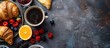 A top-down view of a table featuring a continental breakfast spread with a fresh croissant, berries, jam, black coffee, and a slice of orange. There is space available for text.