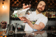 A skilled male bartender confidently pours a martini into a glass at a well-equipped, modern bar, showcasing his expertise and the sophisticated ambience.