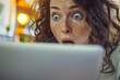 A woman with an expression of shock and amazement looking at her laptop screen, her eyes wide open
