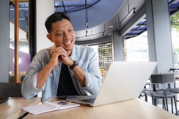 Asian man happy and smile face with blue shirt using laptop and mobile phone in coffee shop, online freelance business