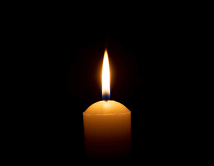 A single burning candle flame or light glowing on a big yellow candle on black or dark background on table in church for Christmas, funeral or memorial service with copy space