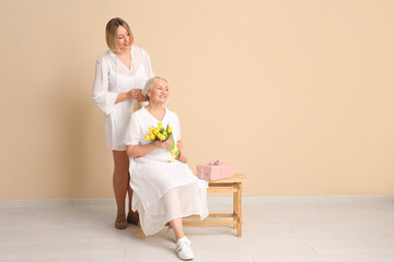 Canvas Print - Mature woman with her daughter, bouquet of tulips and gift box sitting on bench near beige wall. International Women's Day celebration