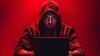   A man in a hoodie sits before a laptop in a dimly lit room, a red light casting an ominous glow from behind