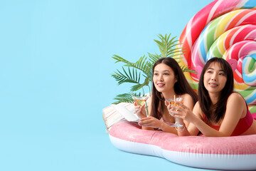 Sticker - Female Asian friends with cocktails lying on swim mattress against blue background