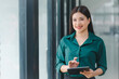 © kenchiro168 - Confident Asian businesswoman smiling and using a digital tablet in a modern office setting, poised for productivity.