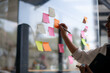 © Wasana - Smiling Asian businesswoman using colorful sticky notes for brainstorming on a glass wall in a modern office.