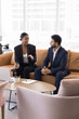© fizkes - Two diverse business professional couple talking in office hall, sitting on couch in coworking space, speaking on break. African manager woman speaking to Indian colleague man. Vertical shot