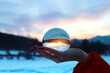 Closeup womans hand holding crystal sphere with scenic cloudy sunset on the background. Winter in mountains.