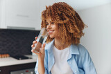 Fototapeta  - Young African American woman enjoying a refreshing drink of water in her stylish kitchen at home