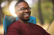 © peopleimages.com - Camping, portrait and black man relax outdoor on vacation in wilderness for peace and wellness in Norway. Person, happy and chair in forest woods for adventure in nature, summer holiday and fresh air