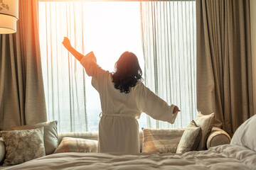 Wall Mural - Hotel relaxation on lazy day with Asian woman waking up from good sleep on bed in weekend morning resting in comfort bedroom looking toward city view, having happy, work-life quality balance lifestyle