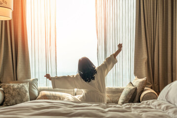 Wall Mural - Hotel relaxation on lazy day with Asian woman waking up from good sleep on bed in weekend morning resting in comfort bedroom looking toward city view, having happy, work-life quality balance lifestyle