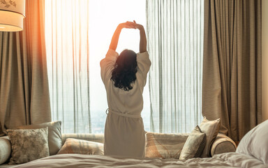 Wall Mural - Hotel relaxation on lazy day with Asian woman waking up from good sleep on bed in weekend morning resting in comfort bedroom looking toward city view, having happy, work-life quality balance lifestyle