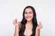 Hopeful young Asian woman in maroon dress crossing fingers on white background