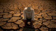 © Nano Photos - Person in a sun hat kneeling on cracked dry earth, suggesting severe drought conditions or possibly examining the soil.