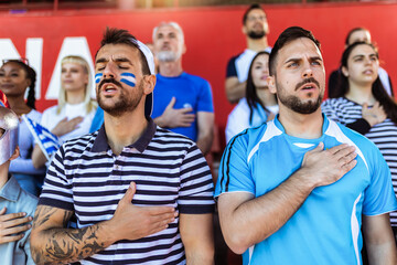 Sport fans singing national anthem of their country at the stadium. Wearing blue and white to support their team.