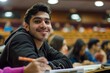 An upbeat young man with a notebook is seated in a university lecture hall, looking at the camera with a friendly smile