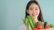 Portrait of a young Asian woman - a happy smiling brunette with long hair, holding a paper bag with vegetables on a cyan background.