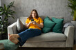 Beautiful Caucasian pleased woman smiling and using cellphone while sitting on sofa in apartment. Phone Communication and comfort concept, full body