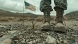 A soldier stands in the desert with his boots next to a small plant. In the background, an American flag is waving.