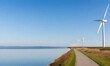 A long path along the shore of a calm (lake, with wind turbines in the distance against a clear blue sky)