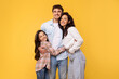 © Home-stock - Healthy family relationship. Loving parents and daughter embracing, standing over yellow studio background and smiling at camera